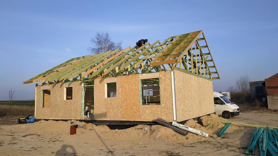 Wznoszenie szkieletu domu z drewnianą konstrukcją dachu i ścianami z płyt OSB, pracownik montuje elementy na dachu, na zewnątrz zaparkowany biały van.