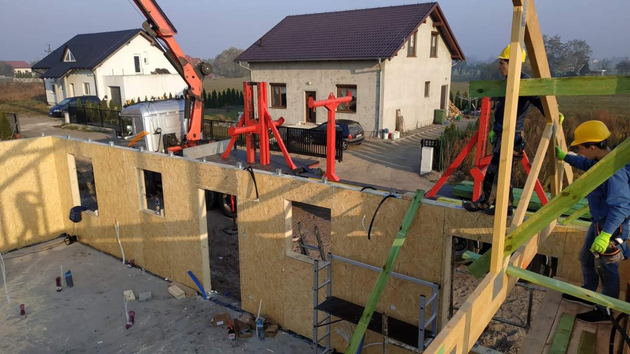 Montaż ścian szkieletowych domu jednorodzinnego z użyciem dźwigu, widoczne elementy konstrukcyjne i dwóch pracowników w kaskach.