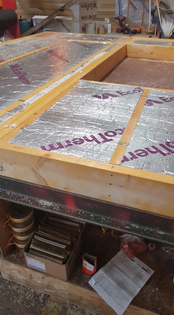 Fragment drewnianej konstrukcji z izolacją termiczną, widoczne materiały budowlane i narzędzia w tle.