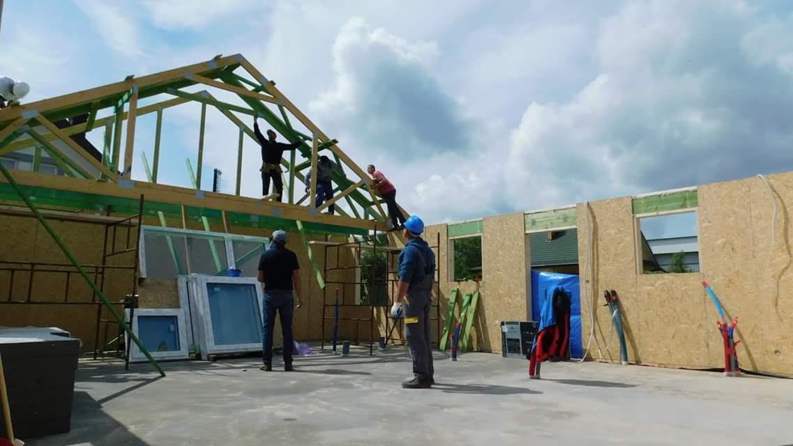 Montaż drewnianej konstrukcji dachu na budynku w technologii szkieletowej; pracownicy na wysokościach, widoczne ściany z płyt OSB, okna i rusztowania.