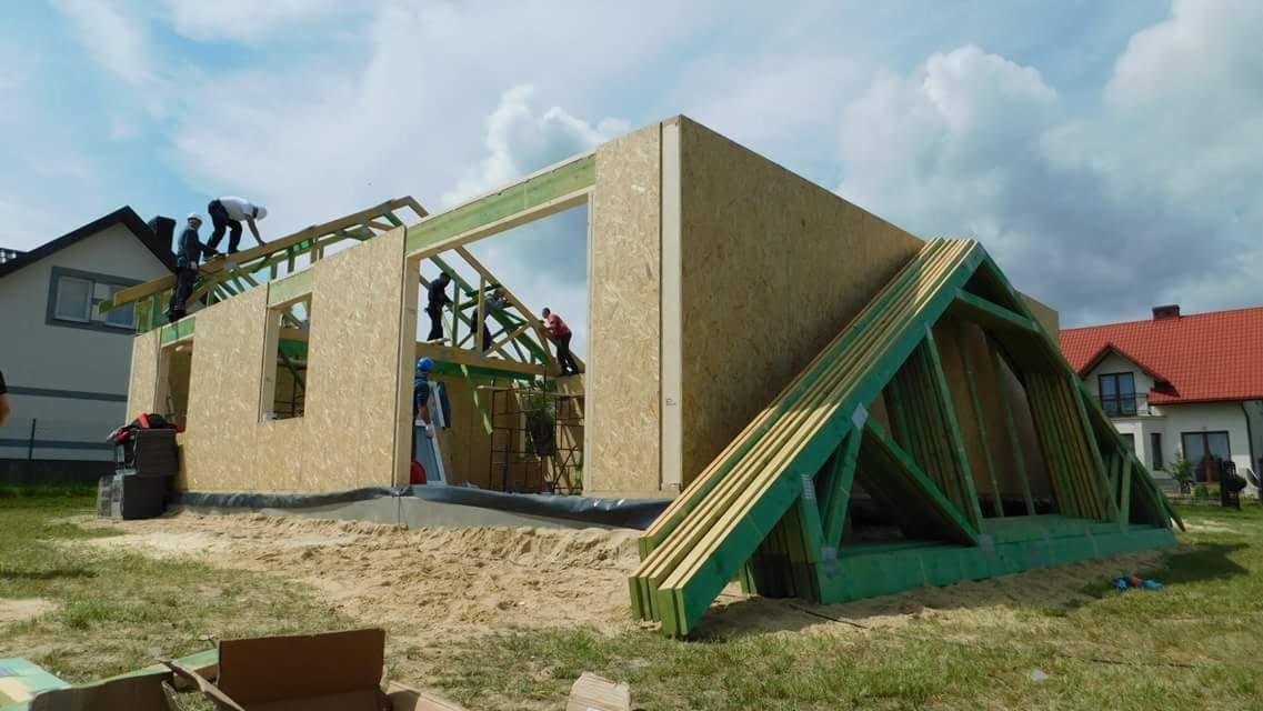 Wznoszenie konstrukcji domu z drewnianych paneli OSB, widoczni pracownicy montujący dach, na pierwszym planie przygotowane elementy więźby dachowej, w tle zabudowa jednorodzinna.