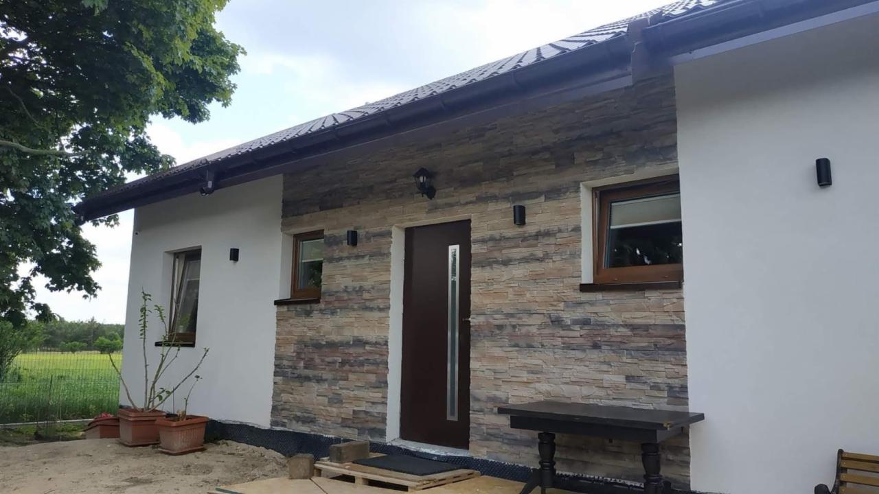 Nowoczesny dom z elewacją z kamienia dekoracyjnego, brązowymi oknami i drzwiami wejściowymi, oświetleniem zewnętrznym i widocznymi elementami izolacji przy ziemi.