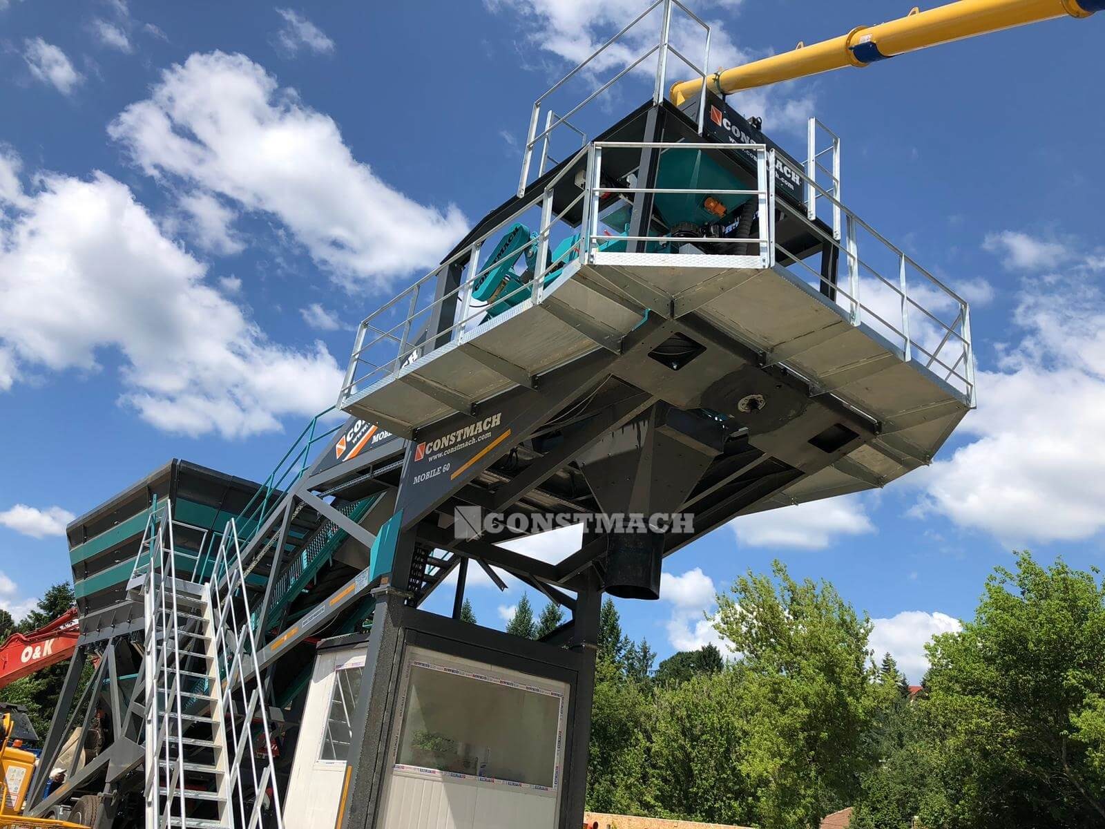 Mobilna betoniarnia CONSTMACH MOBILE 60 z żółtym podajnikiem, widok z dołu na tle błękitnego nieba z chmurami.