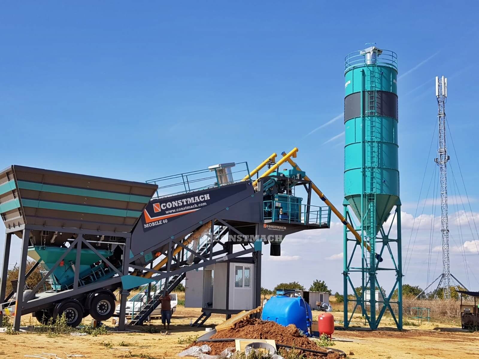 Mobilna betoniarnia MOBILE 60 marki CONSTMACH z silosami na kruszywa i cement, widoczna na tle błękitnego nieba i wieży telekomunikacyjnej.