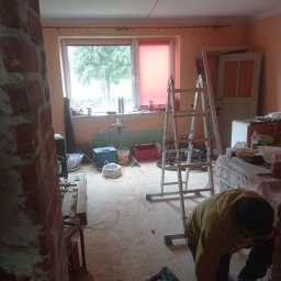 Fazmax Łukasz Zabraniak - Wnętrze w trakcie remontu: odsłonięta cegła, drabina, narzędzia, okno z widokiem na drzewo. Osoba w trakcie pracy, widoczne przewody i sprzęt elektryczny.