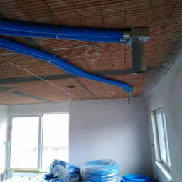 Niebieskie, karbowane rury wentylacyjne zamontowane pod surowym, ceglanym stropem w nowym budynku, z rolkami rur na podłodze i oknami w tle.