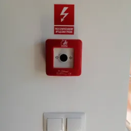 Czerwony, przeciwpożarowy wyłącznik prądu Elektromet z symbolem płomienia, umieszczony na białej ścianie, nad podwójnym włącznikiem światła. Nad wyłącznikiem naklejka z symbolem błyskawicy...