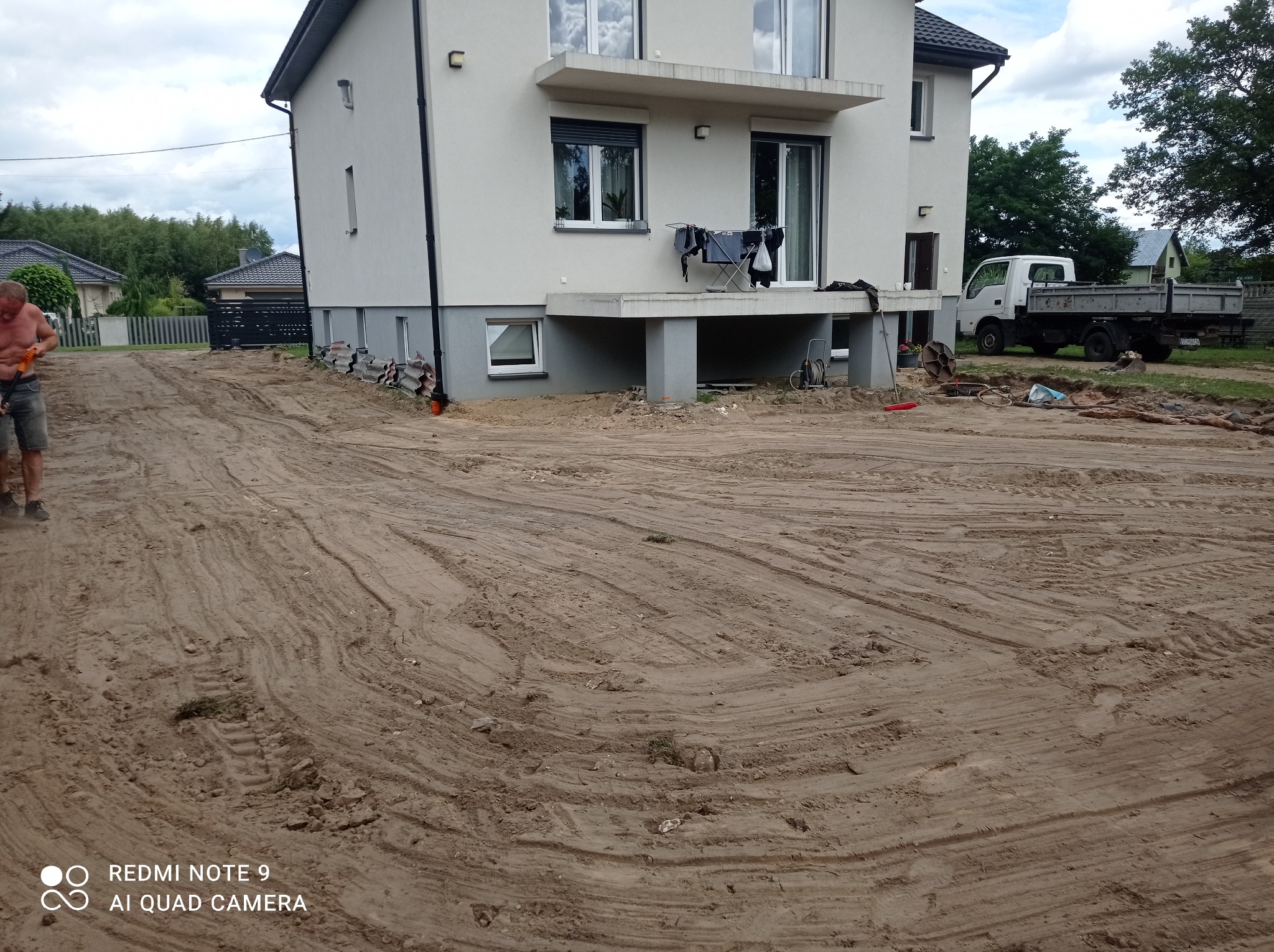 Przygotowanie terenu pod budowę domu jednorodzinnego. Widoczne ślady opon na ziemi, budynek w tle i częściowo widoczna osoba. Prace ziemne w toku.