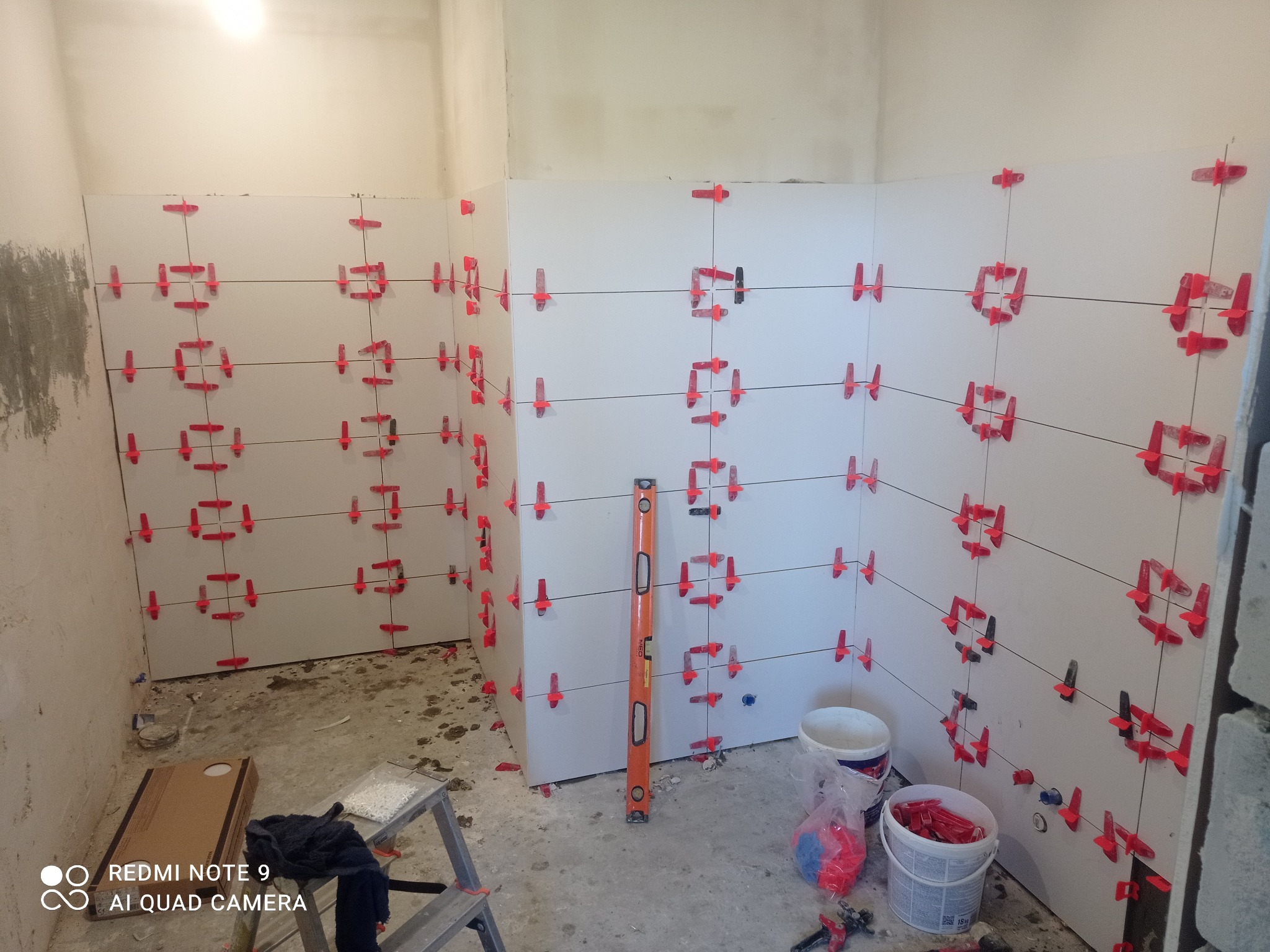 Białe płytki ścienne w trakcie układania, z czerwonymi krzyżykami dystansowymi i poziomicą, w surowym wnętrzu łazienki. Widoczne narzędzia i akcesoria glazurnicze.