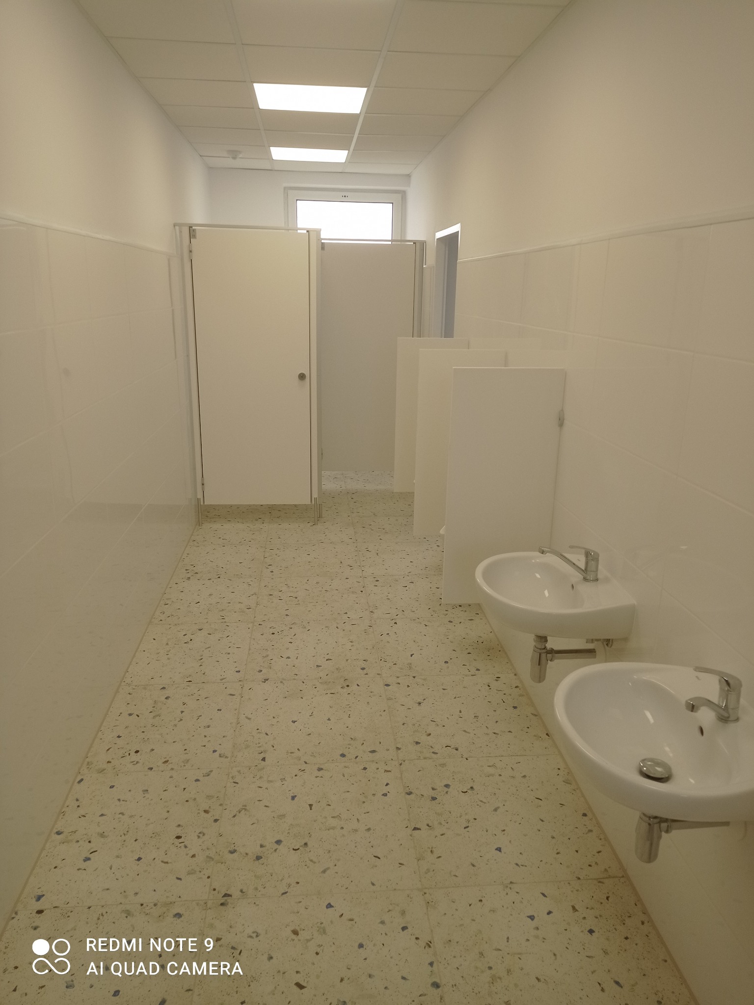 Jasne, wykafelkowane wnętrze publicznej toalety z umywalkami i kabinami WC. Widok z perspektywy wejścia, ukazujący czystość i minimalizm wykończenia.