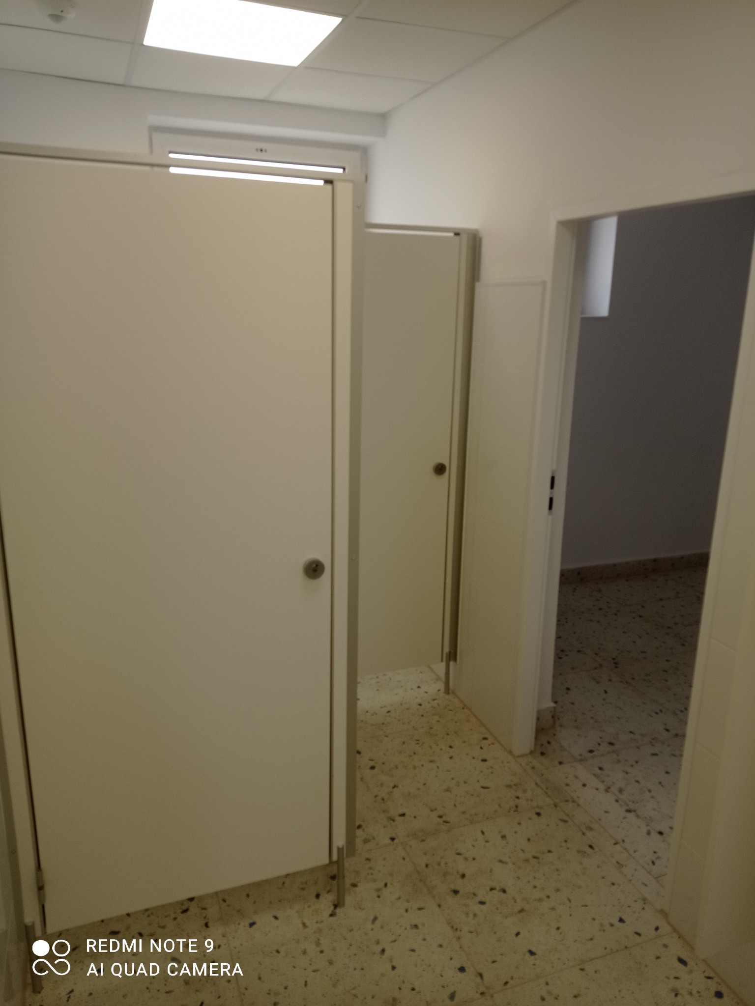 Białe kabiny sanitarne z metalowymi okuciami w jasnym pomieszczeniu z terakotą. Widoczne oświetlenie sufitowe i otwarte drzwi do kolejnych kabin.