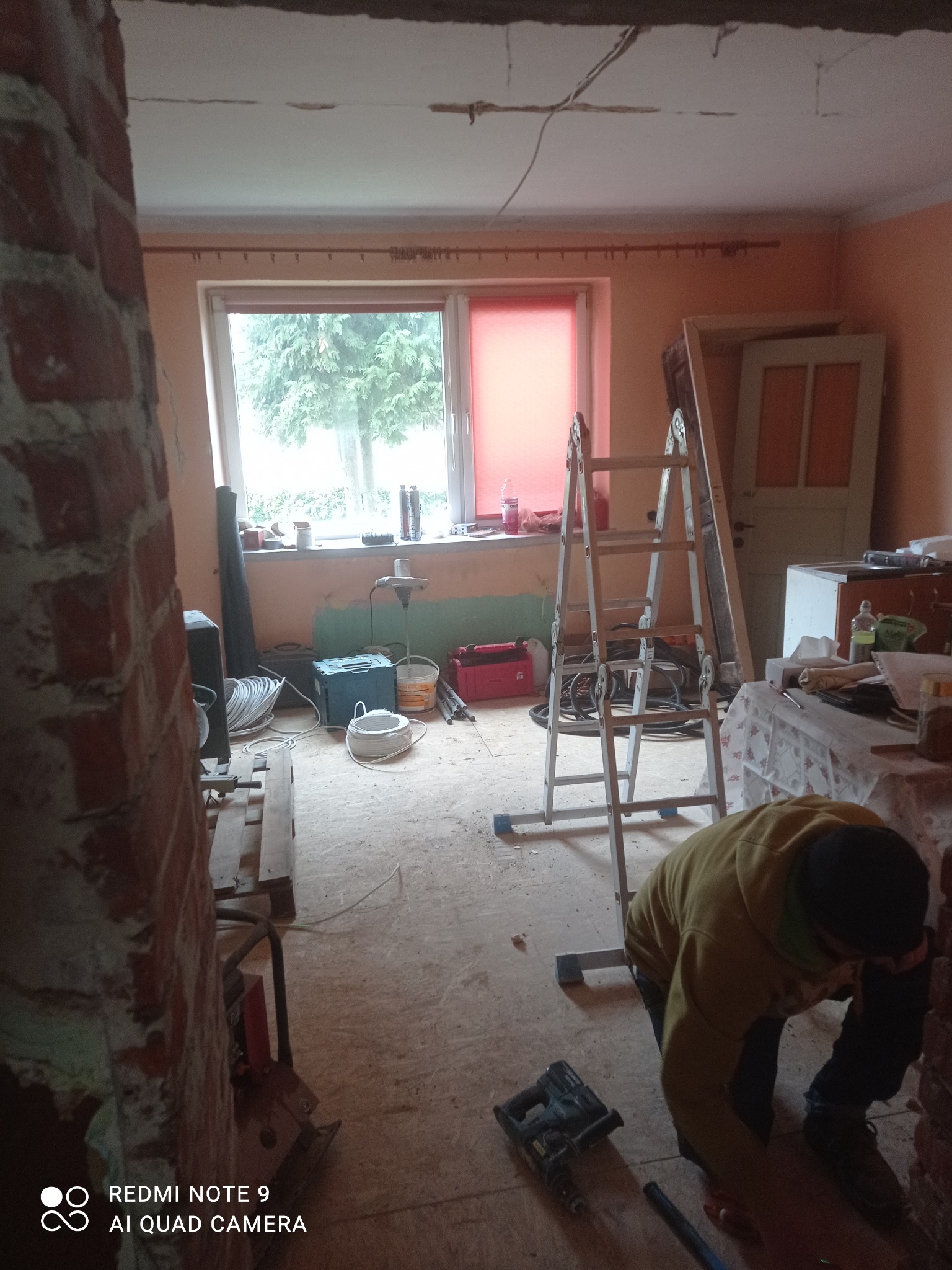 Wnętrze w trakcie remontu: odsłonięta cegła, drabina, narzędzia, okno z widokiem na drzewo. Osoba w trakcie pracy, widoczne przewody i sprzęt elektryczny.
