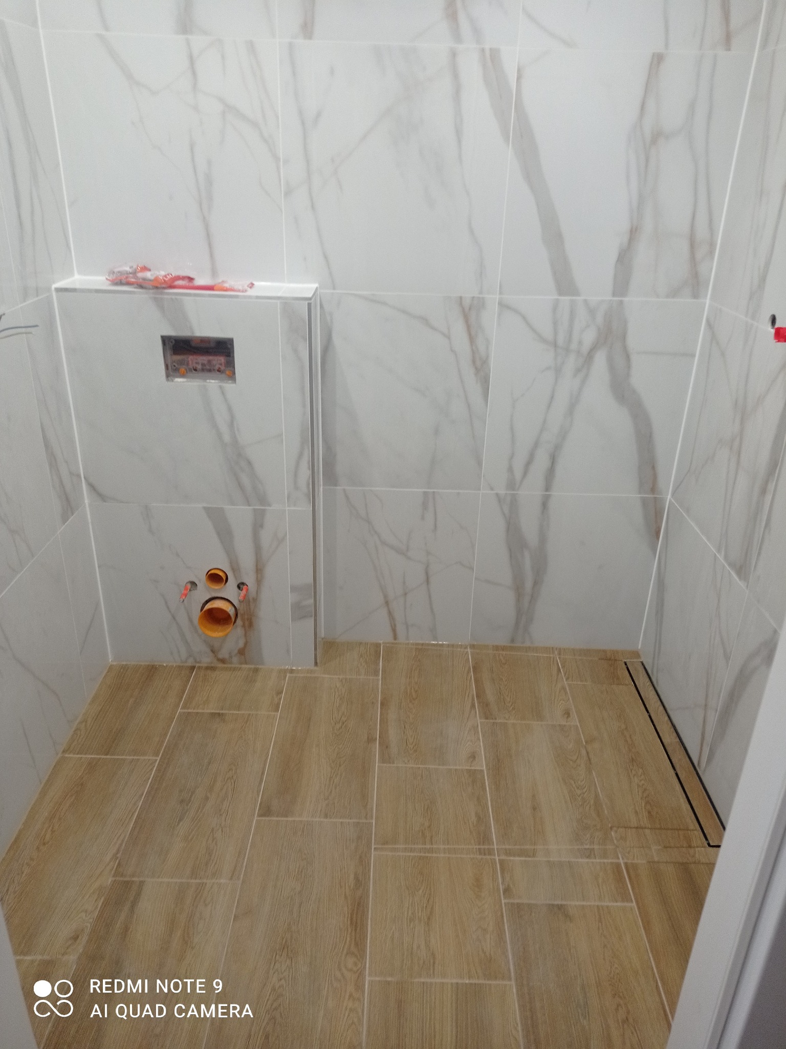 Wykończona łazienka z marmurowymi płytkami ściennymi i drewnopodobną podłogą. Widoczne przyłącza hydrauliczne i odpływ liniowy. Minimalistyczny design.