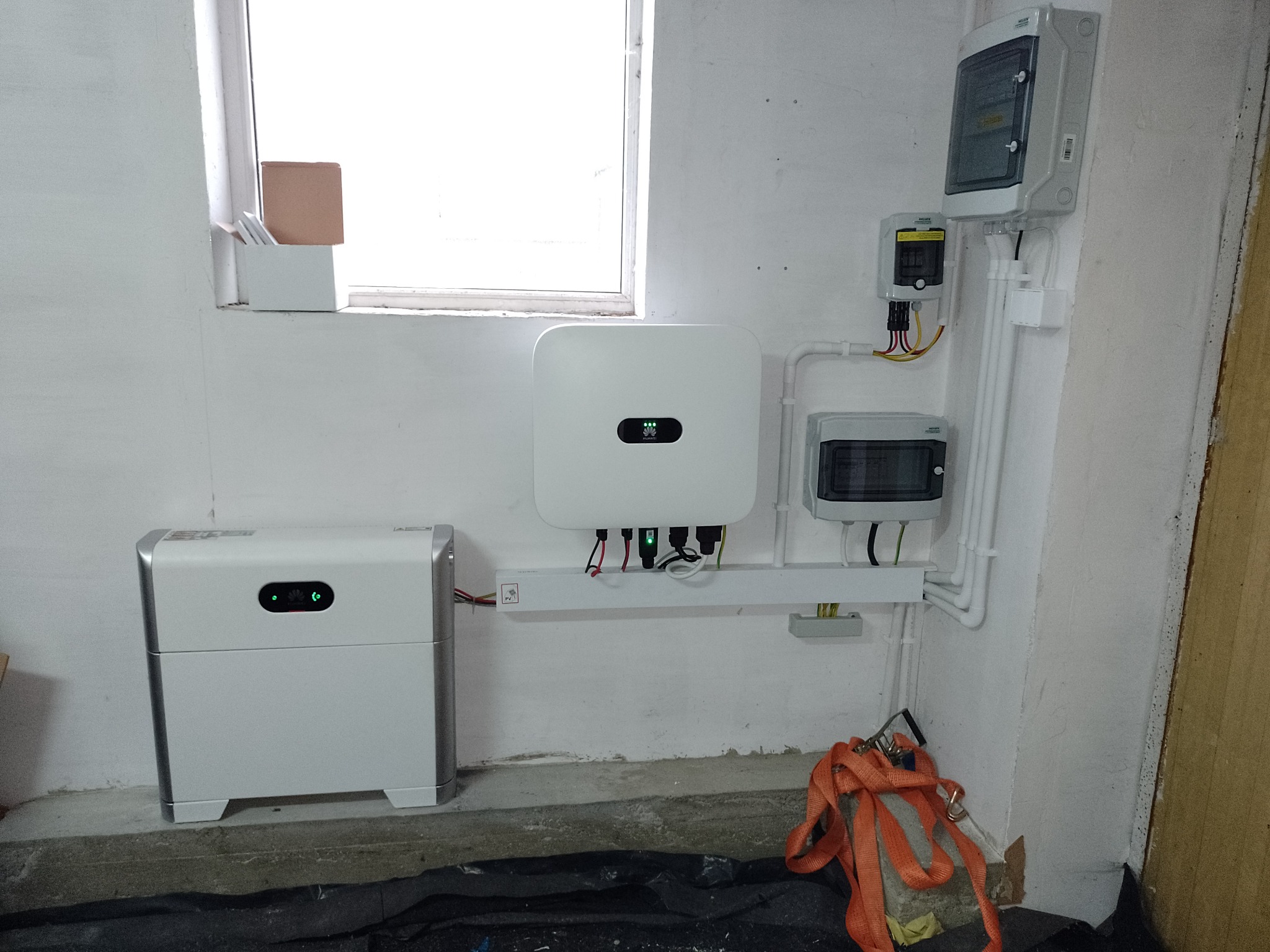 Białe urządzenia elektryczne, w tym falownik i akumulator, zamontowane na ścianie w surowym pomieszczeniu z widocznymi przewodami i skrzynkami rozdzielczymi.