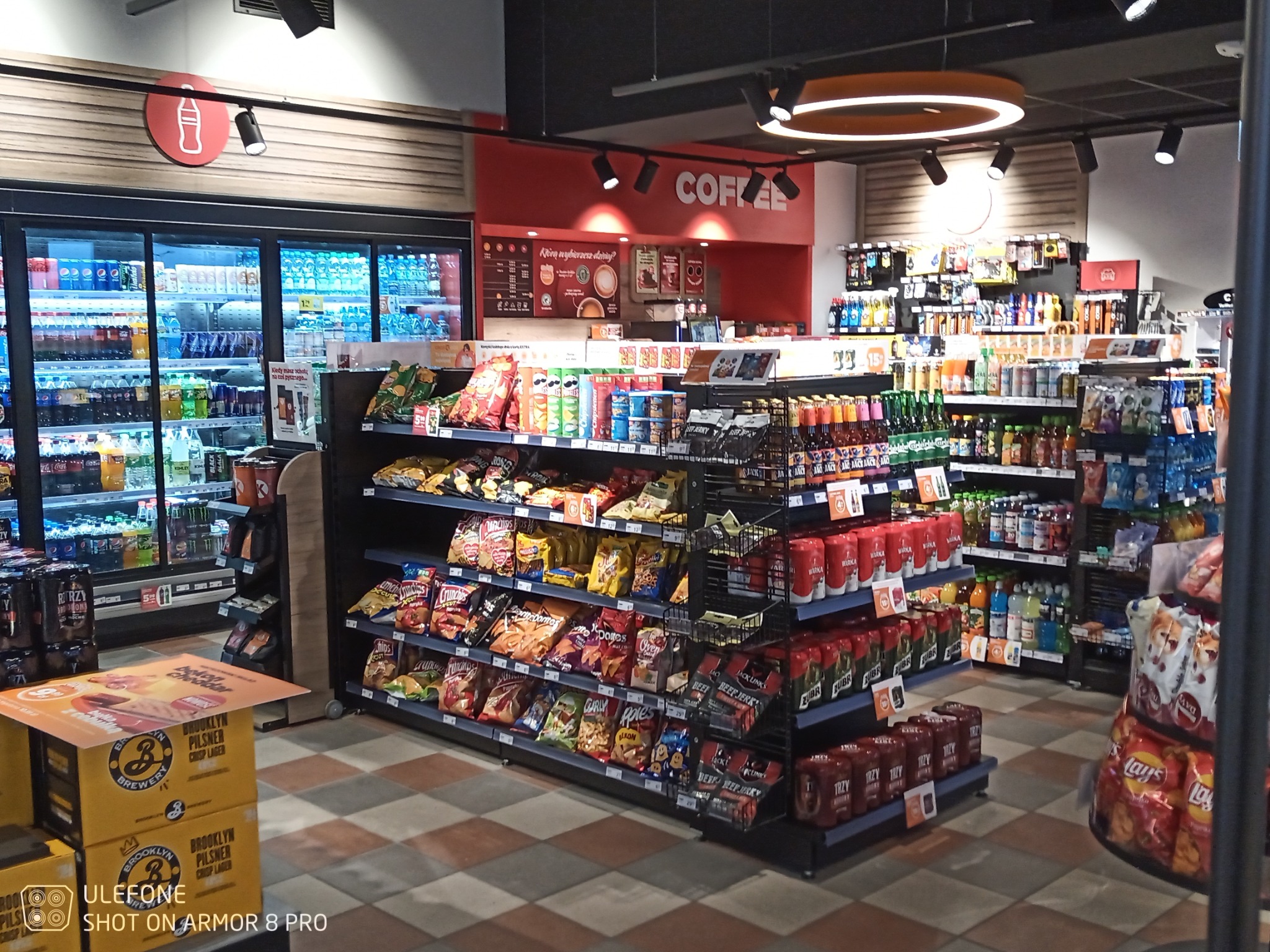 Wnętrze sklepu spożywczego z półkami pełnymi przekąsek, napojów i artykułów spożywczych, oświetlone nowoczesnymi lampami sufitowymi, z widocznym logo Coca-Coli i napisem 'Coffee' w tle