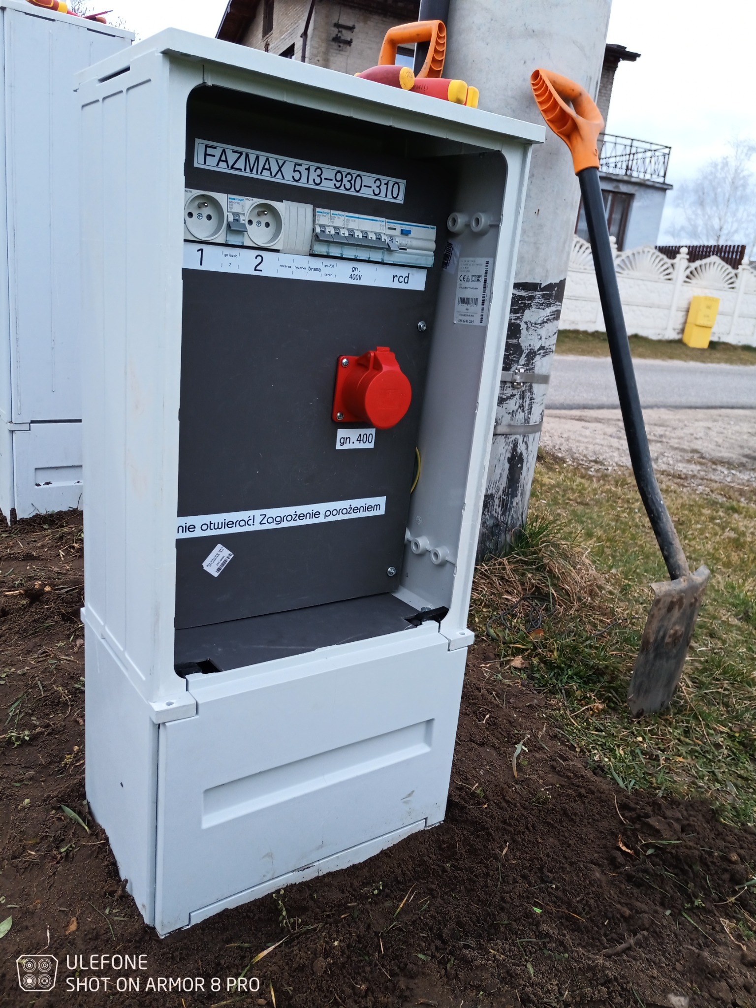 Nowo zainstalowana, otwarta szafka elektryczna FAZMAX z gniazdami, bezpiecznikami i czerwonym gniazdem siłowym gn.400, stojąca na ziemi obok słupa energetycznego, z łopatą opartą o słup.