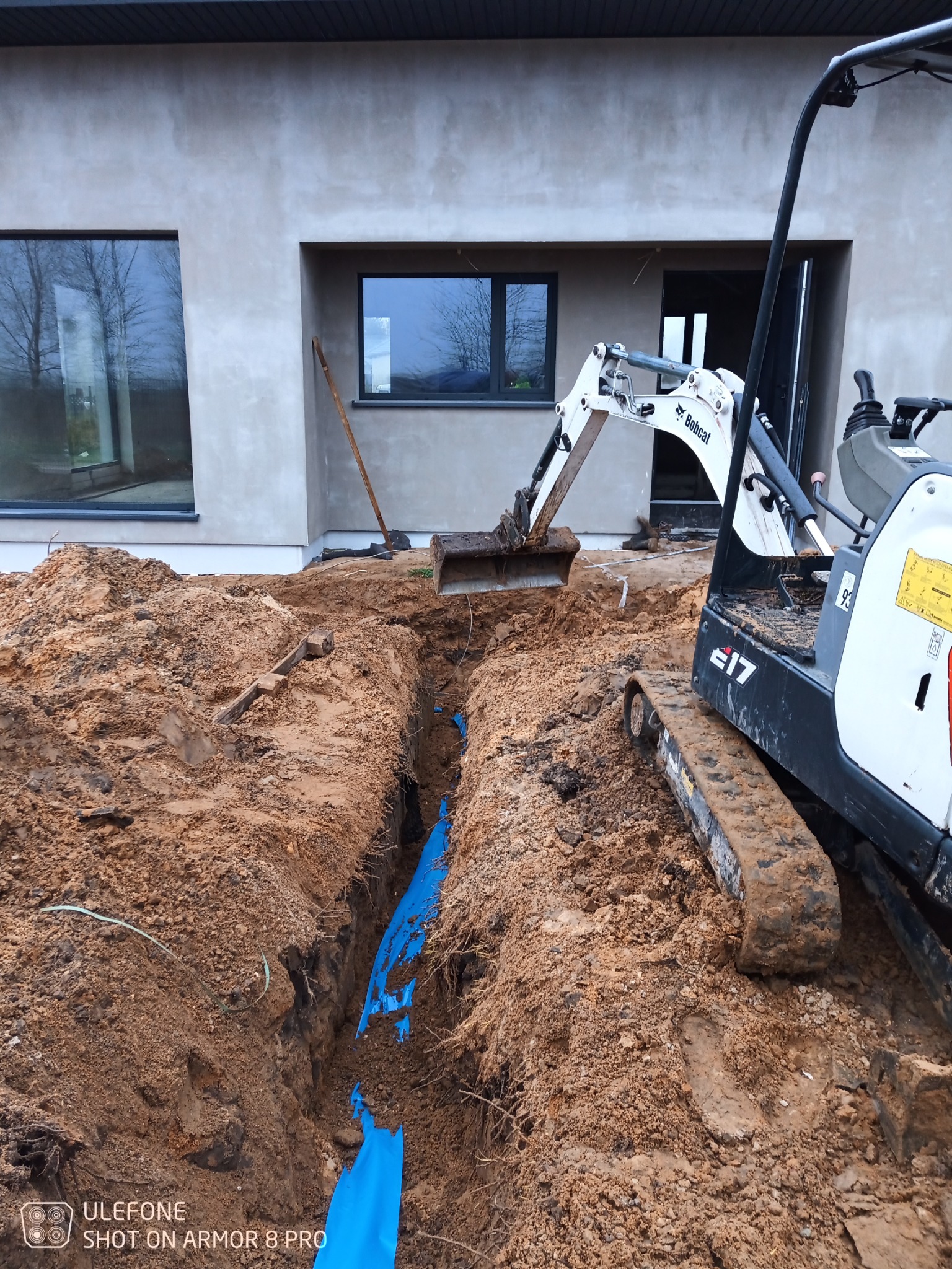 Mini koparka Bobcat E17 obok wykopu z niebieską folią ochronną, na tle surowej elewacji budynku z oknami, w trakcie prac ziemnych.