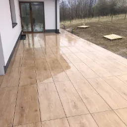 Świeżo ułożona, połyskująca terakota imitująca drewno na tarasie domu jednorodzinnego. Widoczne detale fug i odbicia w mokrej powierzchni. Na zewnątrz widoczny trawnik i drzewa.