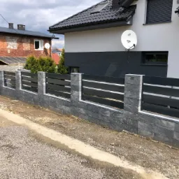 Nowoczesne ogrodzenie z szarej kostki łupanej i poziomych, grafitowych paneli, oddzielające posesję z białym domem i satelitą od starszego, ceglanego budynku.