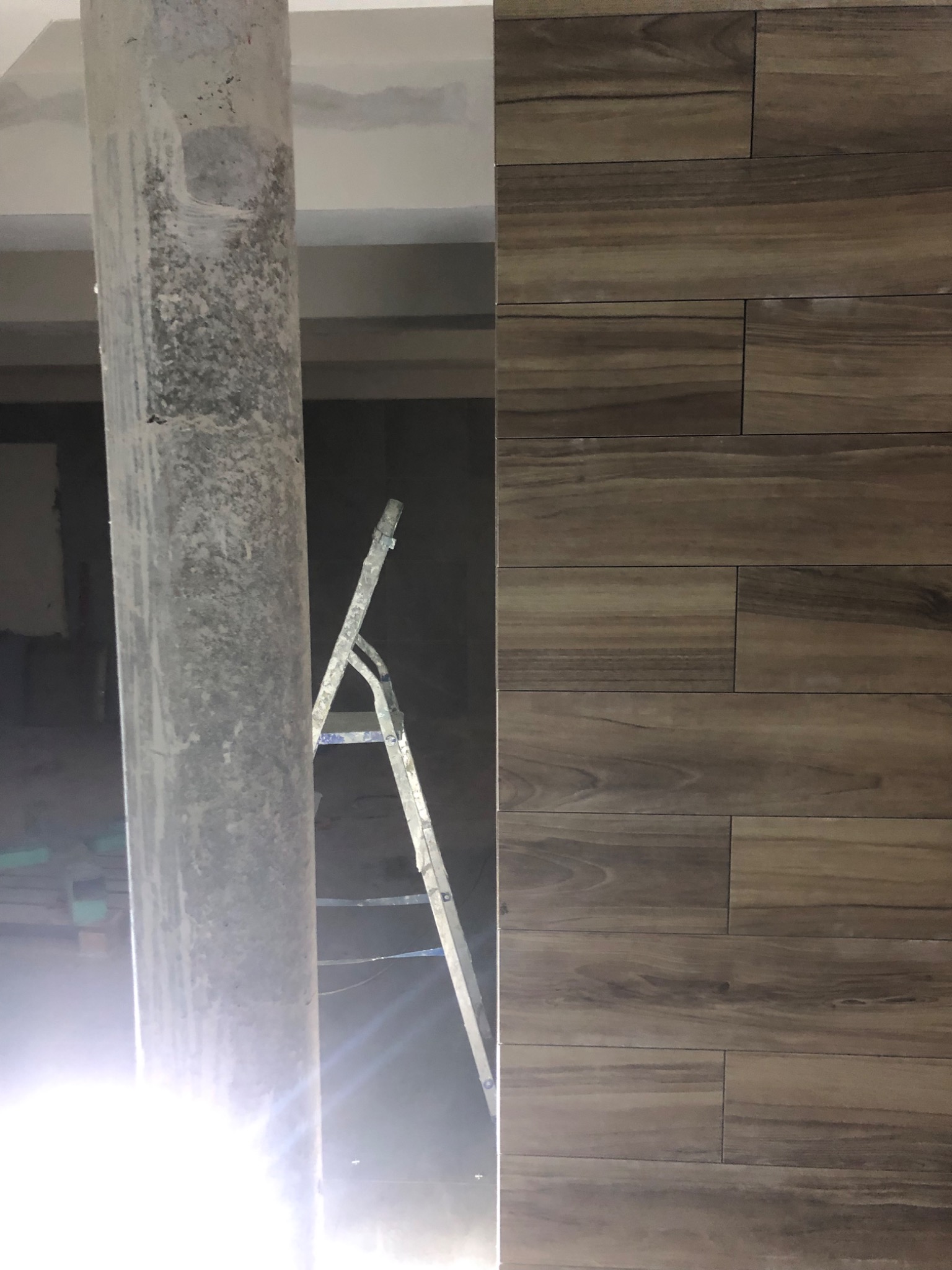 Kolumna w surowym stanie obok ściany wyłożonej brązowymi płytkami imitującymi drewno, drabina w tle.