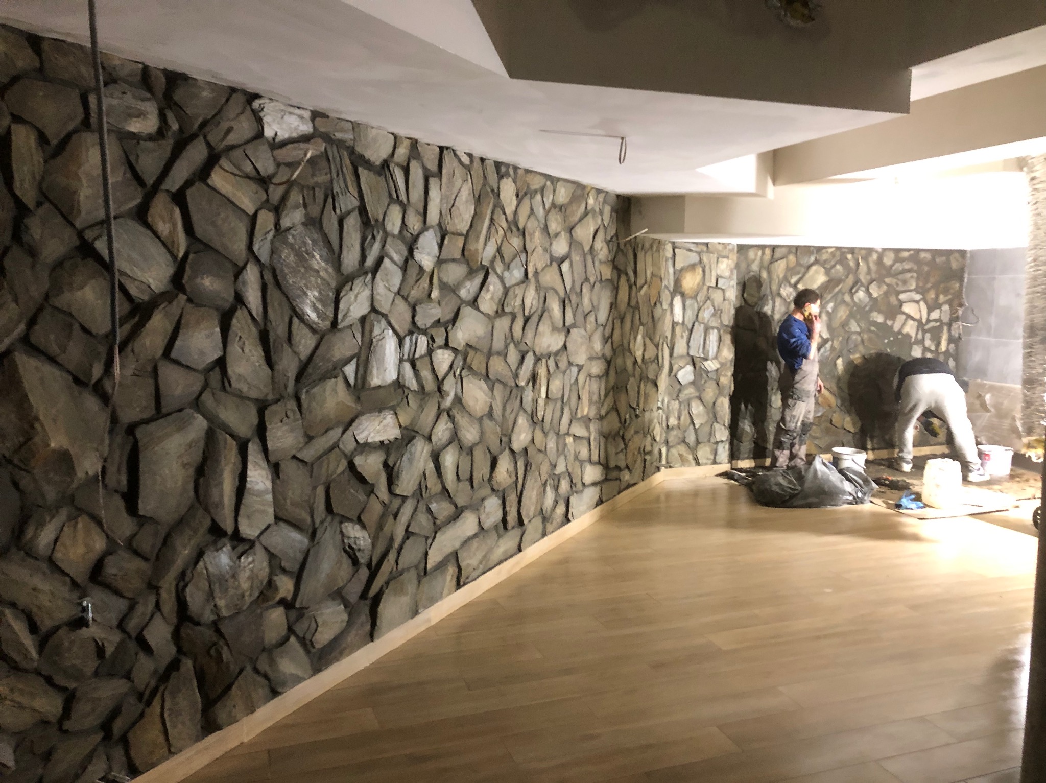 Wnętrze w trakcie prac wykończeniowych: ściana wyłożona kamieniem dekoracyjnym, jasna drewniana podłoga, dwaj pracownicy w tle, jeden rozmawia przez telefon, drugi pracuje nad ścianą.