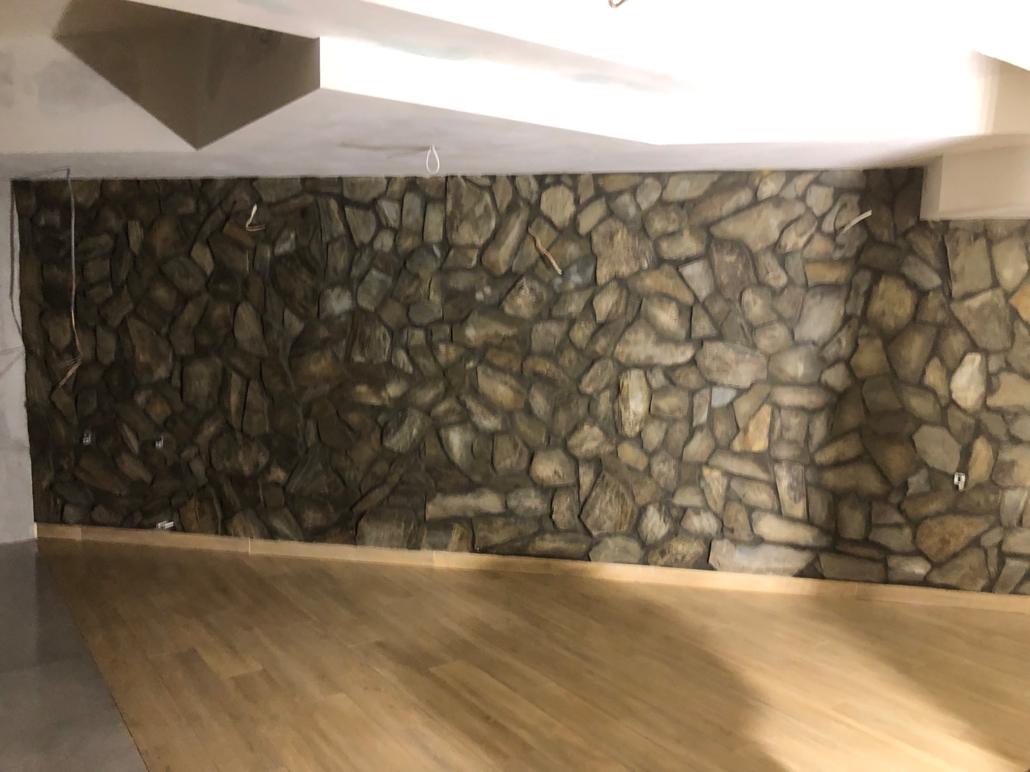 Ściana wyłożona nieregularnymi kamieniami w odcieniach szarości i beżu, kontrastująca z jasną, drewnianą podłogą i białym sufitem z wystającymi przewodami elektrycznymi.