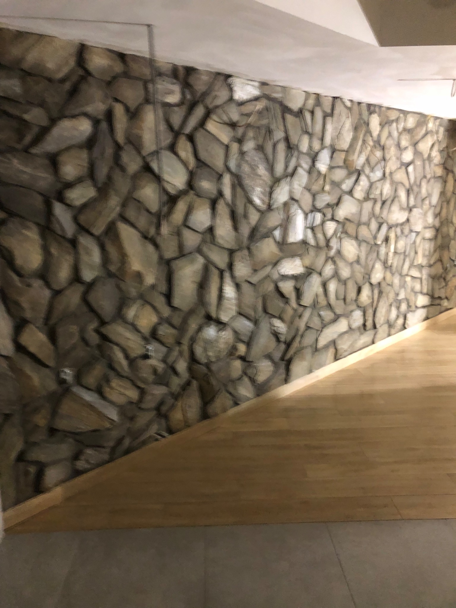 Ściana wykończona kamieniem dekoracyjnym o nieregularnych kształtach, połączona z drewnianą podłogą i szarą podłogą w tle, oświetlona ciepłym światłem.