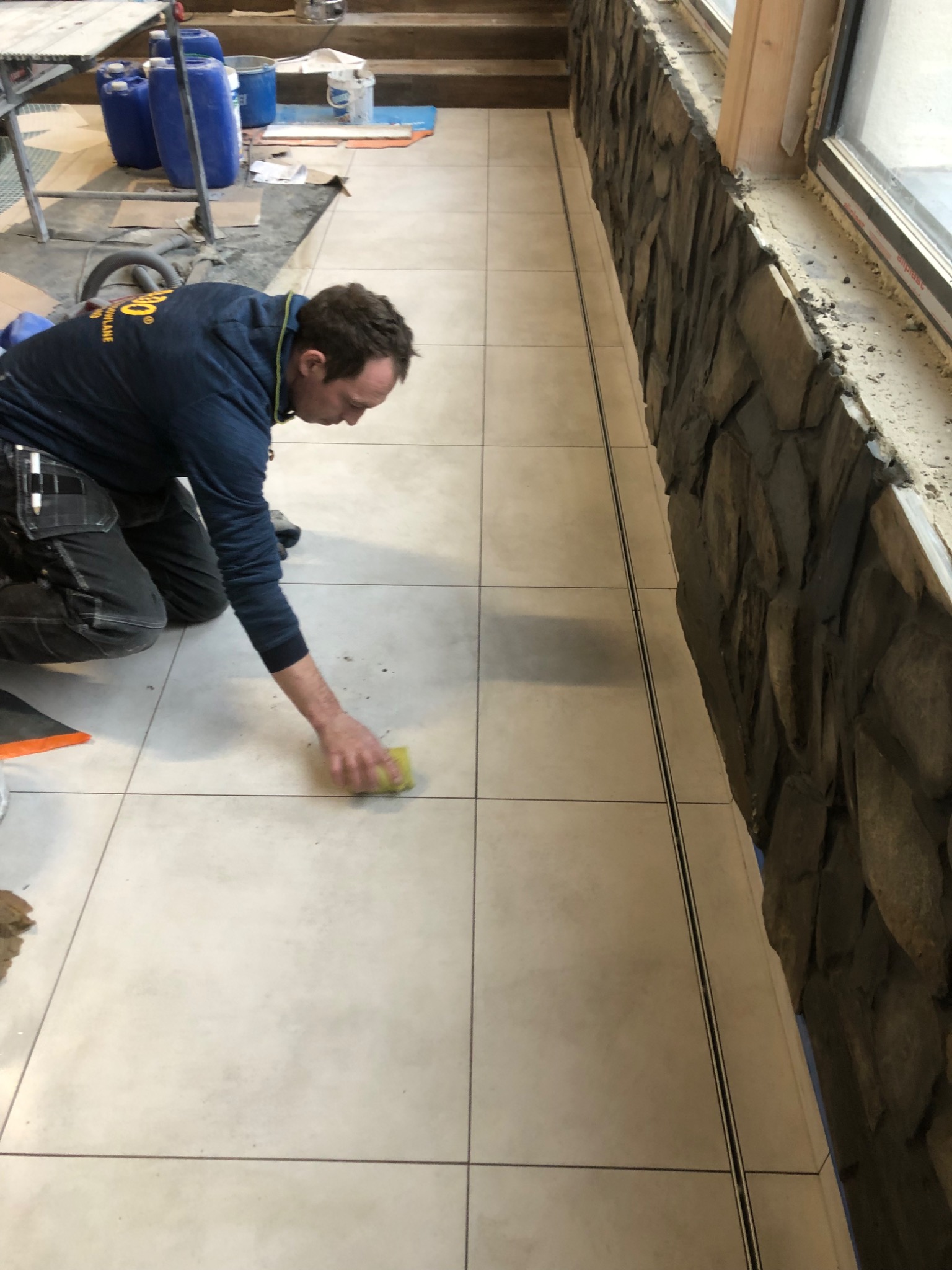 Pracownik na kolanach czyści fugi między jasnymi płytkami podłogowymi żółtą gąbką, obok ściana wykończona kamieniem dekoracyjnym.