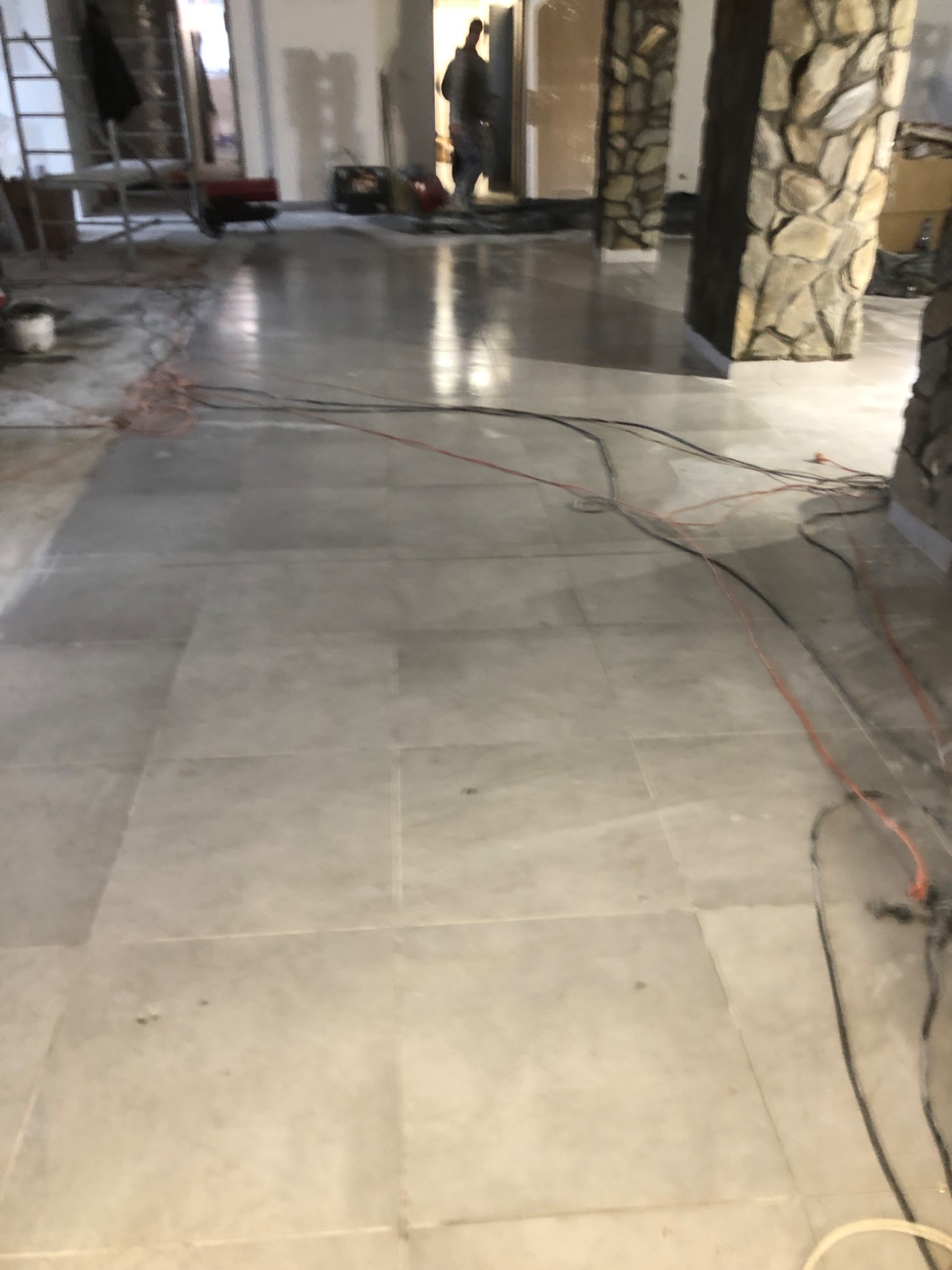 Ułożona, jasnoszara podłoga z płytek w trakcie remontu, widoczne kable, kolumny wykończone kamieniem i sylwetka osoby w tle.