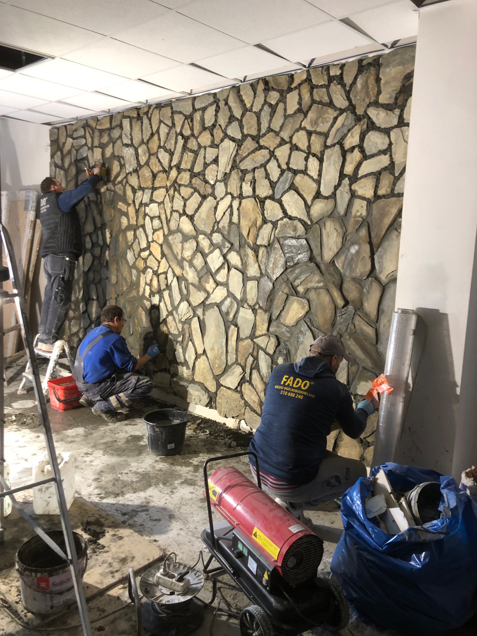 Montaż dekoracyjnej ściany z kamienia przez trzech pracowników; widoczne narzędzia, drabina, grzejnik i worki z odpadami budowlanymi.