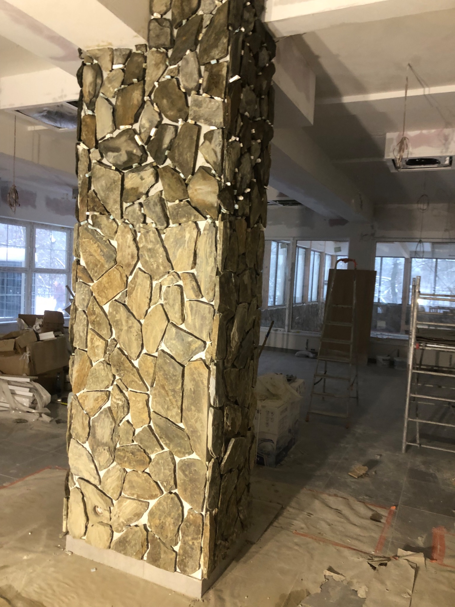 Dekoracyjny filar wyłożony nieregularnymi kamieniami w odcieniach beżu i brązu, widoczny w surowym wnętrzu w trakcie prac wykończeniowych, z widocznymi elementami rusztowania i materiałami...