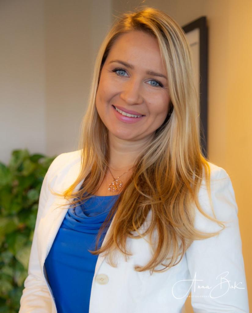 Portret uśmiechniętej kobiety z blond włosami i niebieskimi oczami, ubrana w białą marynarkę i niebieską bluzkę, z widocznym naszyjnikiem w kształcie kwiatu, na rozmytym tle biura.