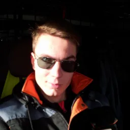 Portret mężczyzny w okularach przeciwsłonecznych i kurtce roboczej z logo firmy 'Partner Techniczny'. Zdjęcie wykonane w ciemnym pomieszczeniu.