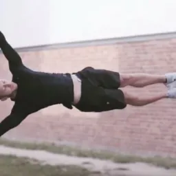 Mężczyzna w czarnej koszulce i spodenkach wykonuje flagę na metalowej drabince przymocowanej do silosów, z ceglanym murem w tle.