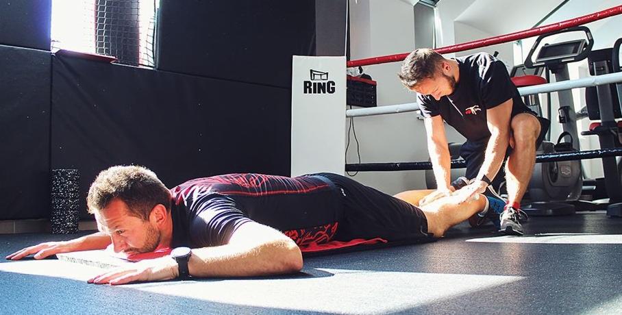 Mężczyzna leżący na brzuchu na macie w siłowni, drugi mężczyzna masuje mu łydkę, w tle ring bokserski i sprzęt do ćwiczeń.
