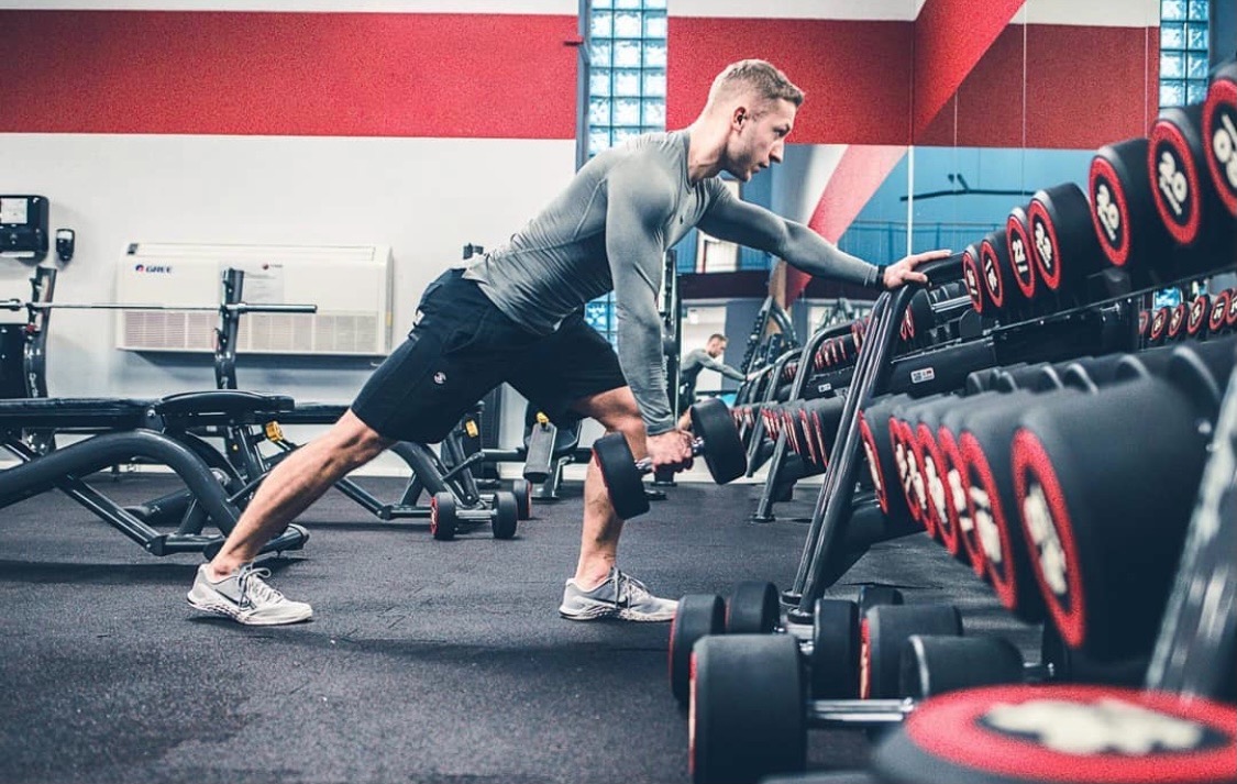 Mężczyzna w szarym stroju sportowym wykonuje wiosłowanie hantlą w oparciu o stojak z hantlami w siłowni.