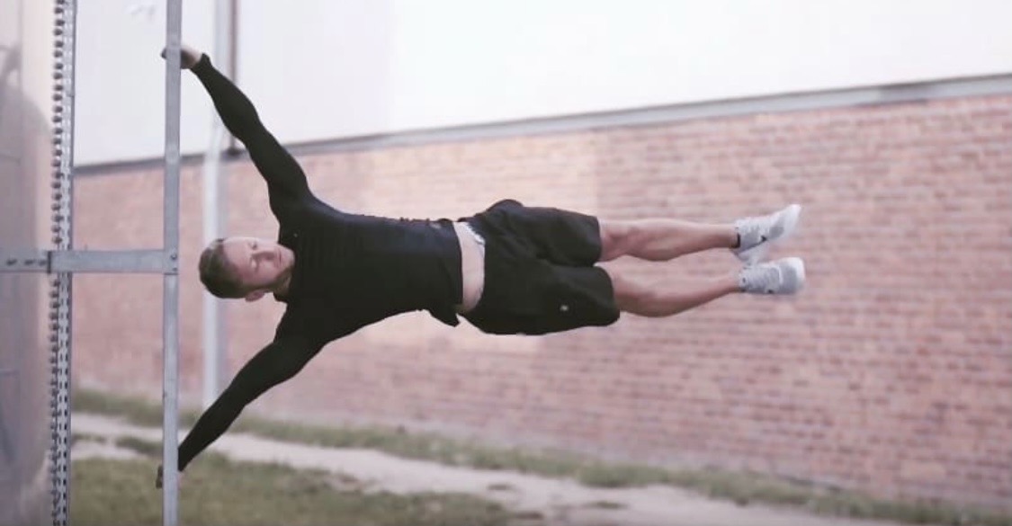 Mężczyzna w czarnej koszulce i spodenkach wykonuje flagę na metalowej drabince przymocowanej do silosów, z ceglanym murem w tle.