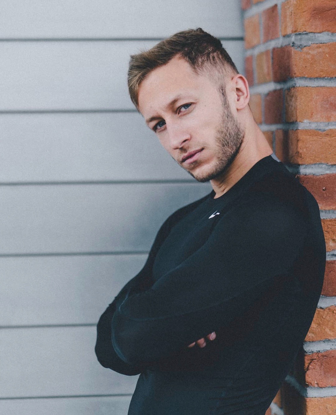 Młody mężczyzna w czarnej koszulce z długim rękawem, oparty o ścianę z cegły i szare panele, z założonymi rękami i intensywnym spojrzeniem.