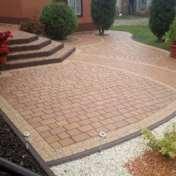 Ułożona kostka brukowa w odcieniach brązu i beżu z obramowaniem z drobnego żwiru, prowadząca do schodów wejściowych z donicami z czerwonymi kwiatami i dalej do ogrodu z krzewami i trawą.