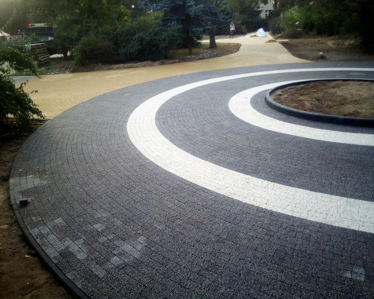 Okrągły podjazd wyłożony ciemnoszarą kostką z białymi pasami, obrzeże z kostki, w tle żółta kostka i zieleń.