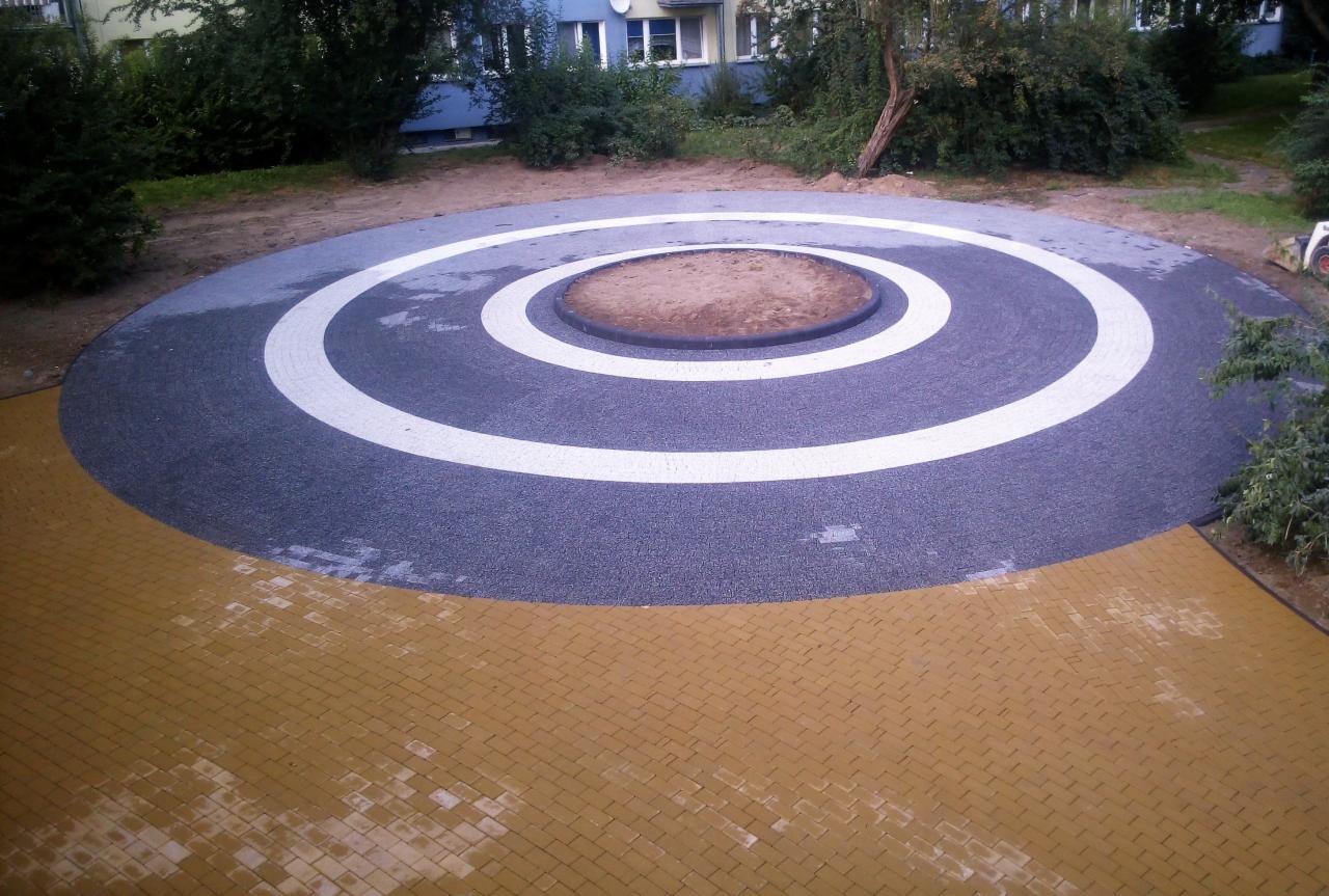 Okrągły plac zabaw z kostki brukowej w kolorze szarym z białymi okręgami, otoczony żółtą kostką brukową. Na środku piaskownica.