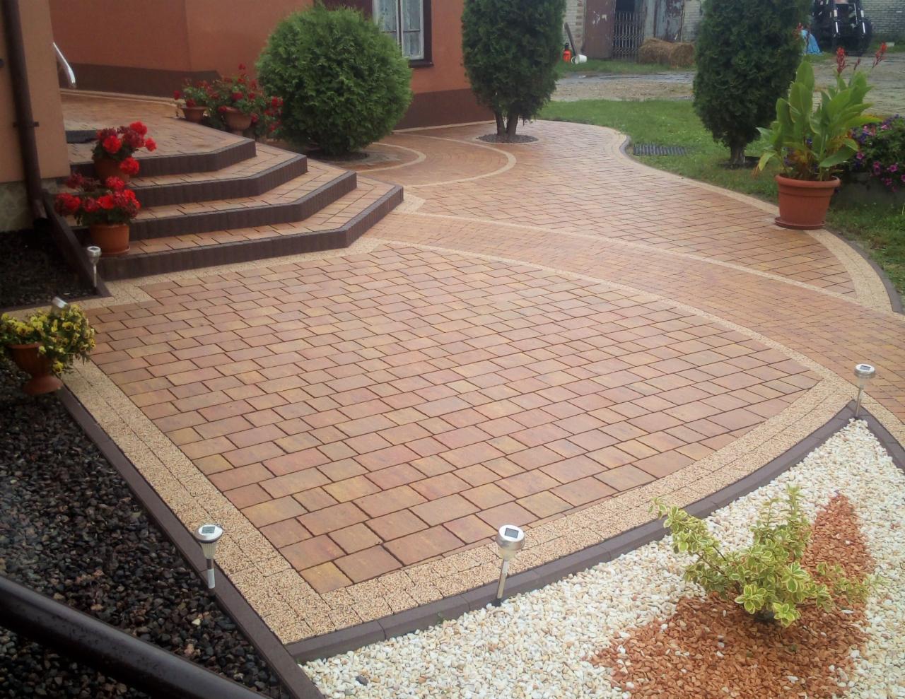Ułożona kostka brukowa w odcieniach brązu i beżu z obramowaniem z drobnego żwiru, prowadząca do schodów wejściowych z donicami z czerwonymi kwiatami i dalej do ogrodu z krzewami i trawą.