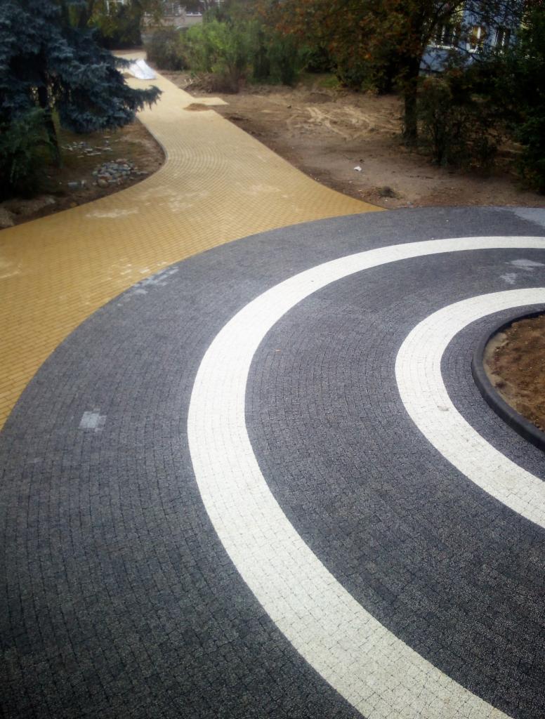 Zakrzywiony chodnik z szarej kostki granitowej z białymi pasami, przechodzący w ścieżkę z żółtej kostki brukowej, otoczenie z ziemią i zielenią.