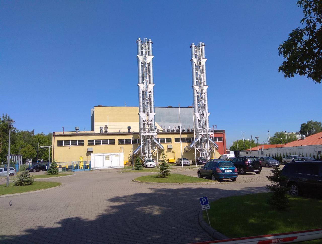 Dwa wysokie, metalowe kominy przemysłowe z konstrukcją wsporczą, umiejscowione przy budynku elektrociepłowni z parkingiem i zielenią w słoneczny dzień.