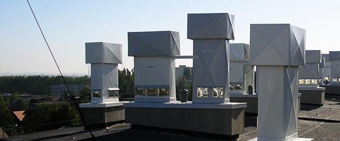 Szereg nowoczesnych, srebrnych kominów wentylacyjnych z kwadratowymi zakończeniami na dachu budynku w Rzeszowie, widok z bliska, błękitne niebo w tle.