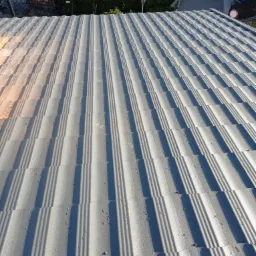 Nowo położony, szary dach z blachy trapezowej, widoczne krople wody i cienie, w tle zieleń i fragmenty budynków.