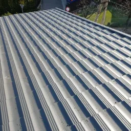 Nowy, szary dach z blachodachówki modułowej, widok z góry z perspektywą na ulicę i domy w tle, krople wody na powierzchni.