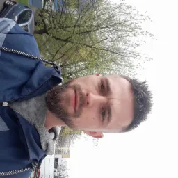 Portret brodatego mężczyzny w niebiesko-szarej kurtce na tle drzewa i budynku. Zdjęcie z dołu, obrócone o 90 stopni.