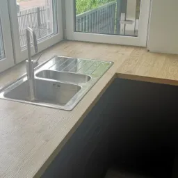 Kuchenny blat drewniany z dwukomorowym zlewem stalowym pod oknem, widok na ulicę z szarą zabudową i samochodem