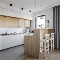 Nowoczesna kuchnia z białymi szafkami i drewnianymi akcentami, wyspą z dwoma białymi krzesłami barowymi, szarymi zasłonami i podłogą łączącą szare płytki i drewniane panele.